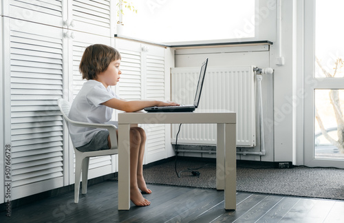 Boy sits at the table, uses laptop, typing on a keyboard, looks at the screen. Child does homework lesson, plays video game, study IT course. Home distance online education concept. Smart technology photo