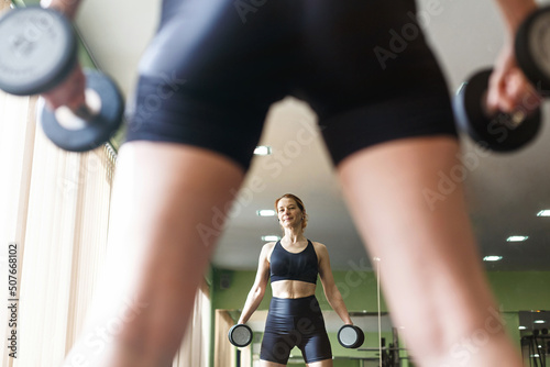 Adult athletic woman in black sportswear performs strength training in the gym. Girl lifts heavy weights on the background of sports equipment. Health, sports lifestyle. View from the back next mirror