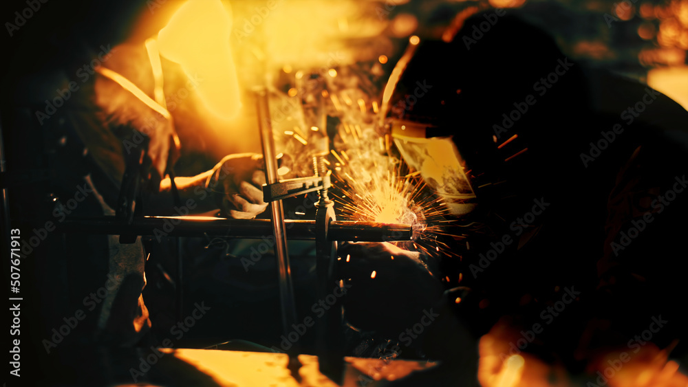 Close up of workers in a welding masks and protective gloves using welding machine in the dark workshop with bright sparks flying into the sides. Stock footage. Workers with professional equipment.