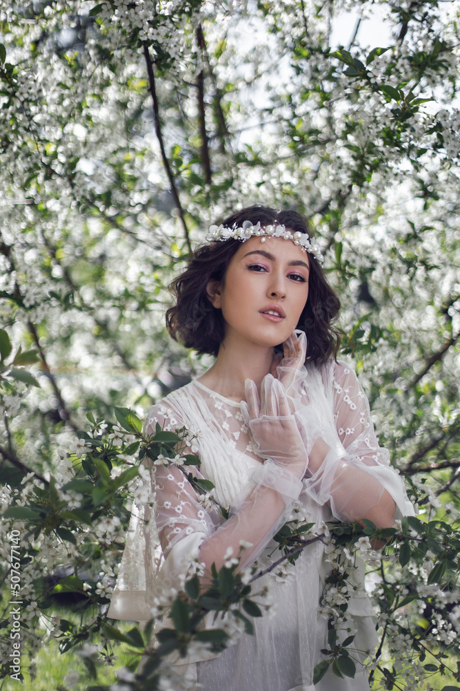portrait of a young beautiful woman in white clothes standing next to a blooming cherry tree in spring. a wreath on the head. portrait of a young beautiful woman in white clothes standing next to a blooming cherry tree in spring. a wreath on the head.