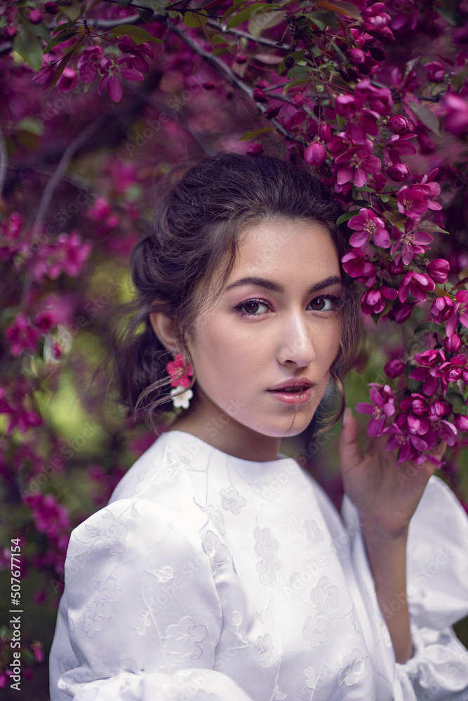 Fototapeta premium portrait sexy young woman in a white dress and purple tights stands by the blooming purple cherry trees in spring.