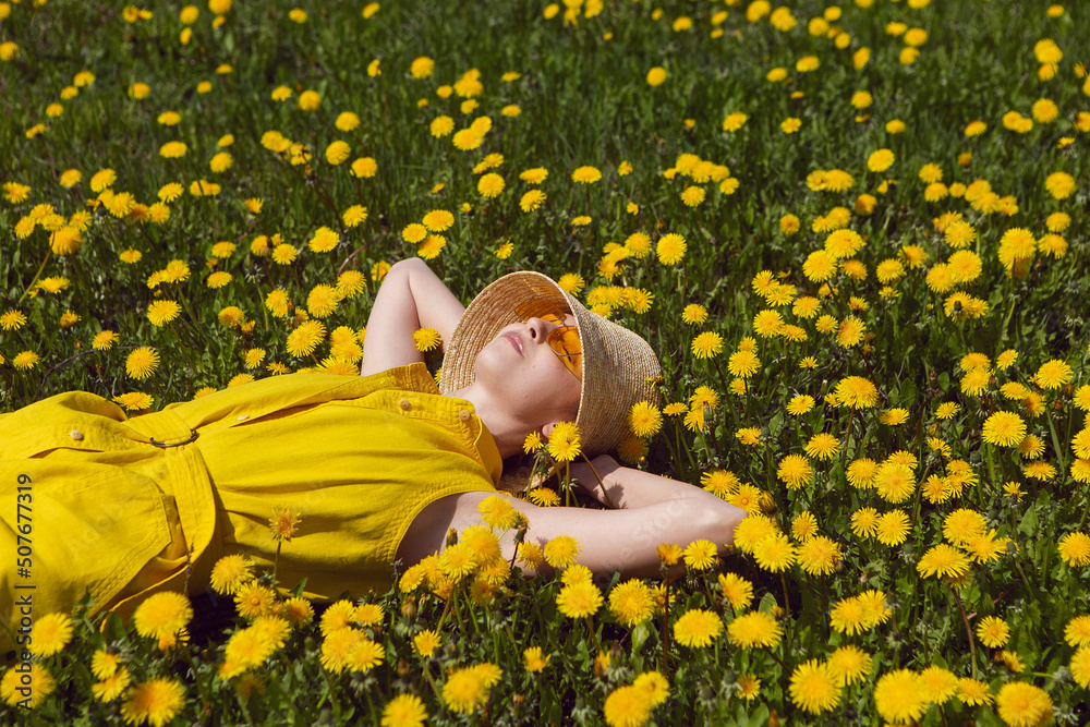 Fototapeta premium woman in a yellow dress, hat and yellow glasses lies on a field of dandelions in summer.