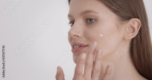Beauty skin care. Attractive caucasian woman with bare shoulders standing in studio and applying face cream with fingers. Concept of skin care and beauty treatment.