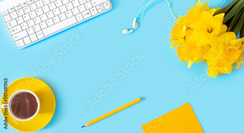 Woman's modern workspace. Keyboard, and office supplies on blue table. Bouquet of delicate beautiful yellow daffodils and cup of tea for holiday.