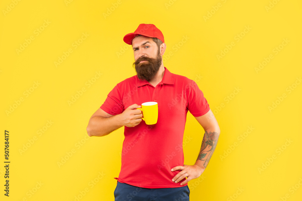 Serious bearded man in casual red cap and tshirt holding mug yellow background, coffee Serious bearded man in casual red cap and tshirt holding mug yellow background, coffee