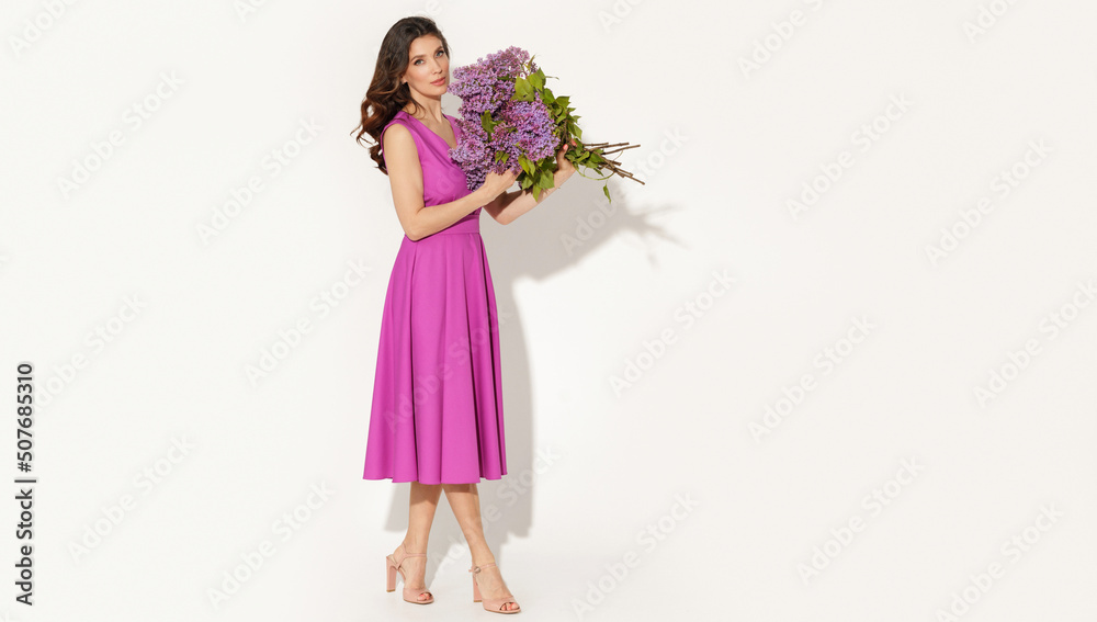Full-length studio fashion portrait of elegant woman wearing purple dress and holding lilac brunches against white wall. International women’s day, Mother’s day and spring concept.