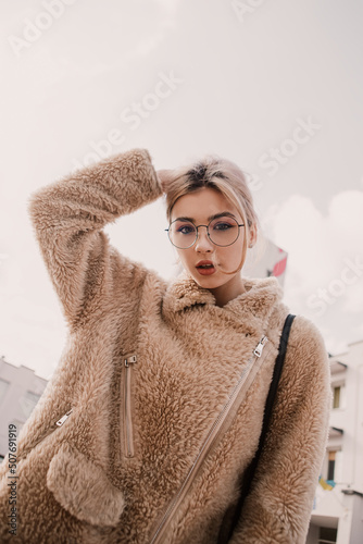 Vertical portrait of young hipster woman wear glasses posing looking at the camera. Blond lady dressed sheepskin coat fur jacket walks oudoors street.
