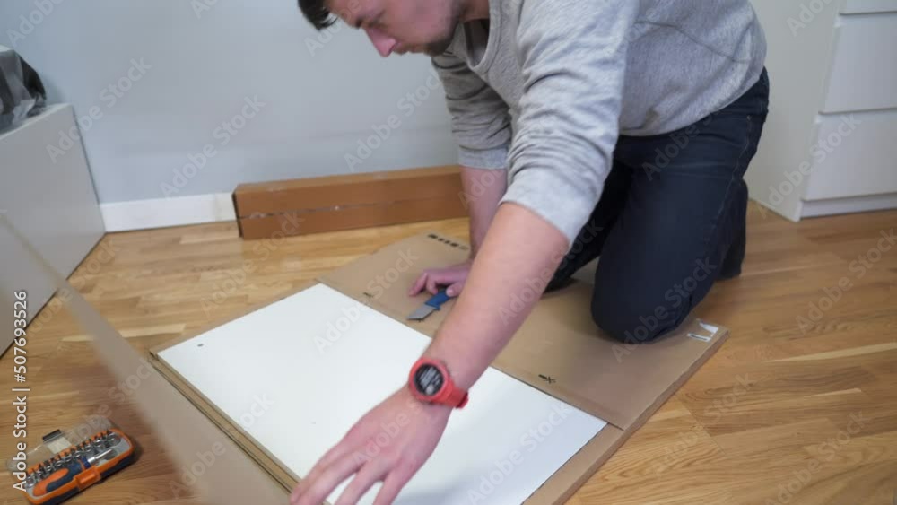The buyer uncovers white table to assemble on the floor of a cardboard box at home. A man bought home furniture and opens the box. New white furniture after relocation. Packaging with new furniture The buyer uncovers white table to assemble on the floor of a cardboard box at home. A man bought home furniture and opens the box. New white furniture after relocation. Packaging with new furniture