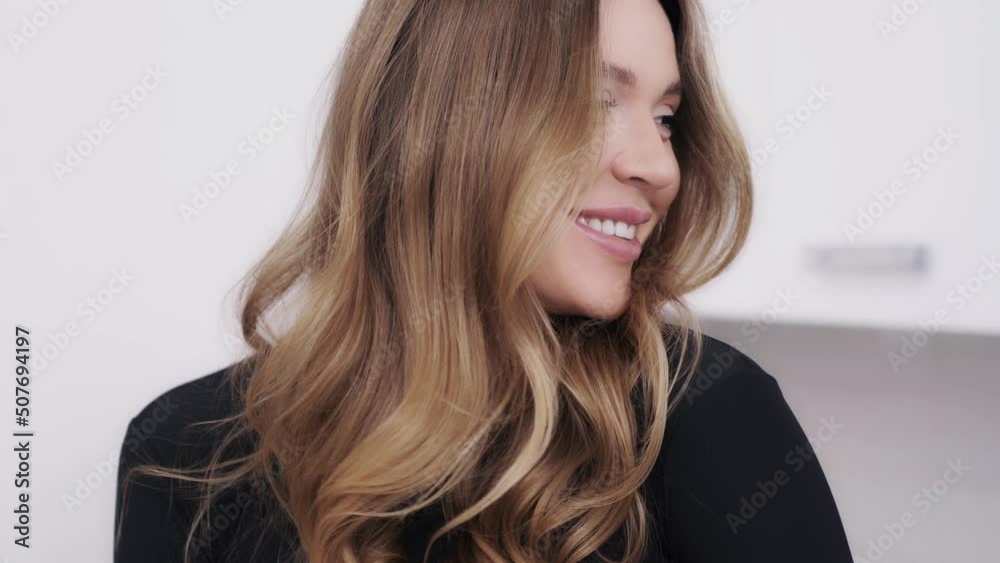 Close-up indoor portrait of a beautiful young woman with dark blond hair looking into the camera with a big smile. Charming girl touching hair being a bit flirty and coquettish
