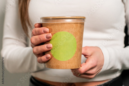 Cardboard disposable cup with plastic lid and light green sticker. The girl holds in his hands, advertises, disposable tableware with a green logo. The concept of disposable tableware. Ecological ware