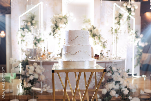 The wedding cake. White, two-tier, decorated with gold. On a delicate light background and a golden stand. Side view.
