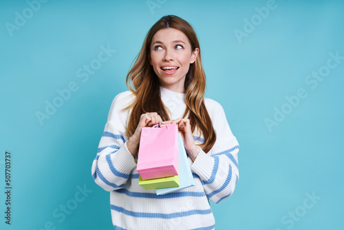 Thoughtful young woman carrying little shopping bags while standing against blue background
