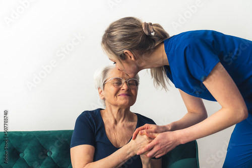 Female MD in her 20s wearing blue scrubs leaning towards an elderly woman sitting on a sofa, kissing her in her forehead. Indoor shot. High quality photo