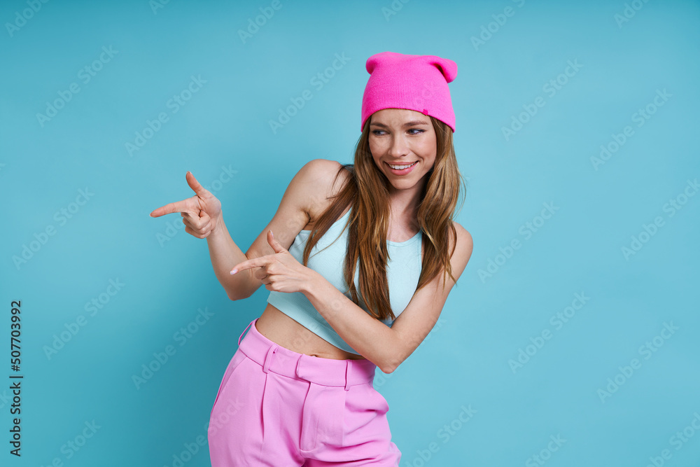 Attractive young woman in pink hat smiling and pointing away while standing against blue background Attractive young woman in pink hat smiling and pointing away while standing against blue background