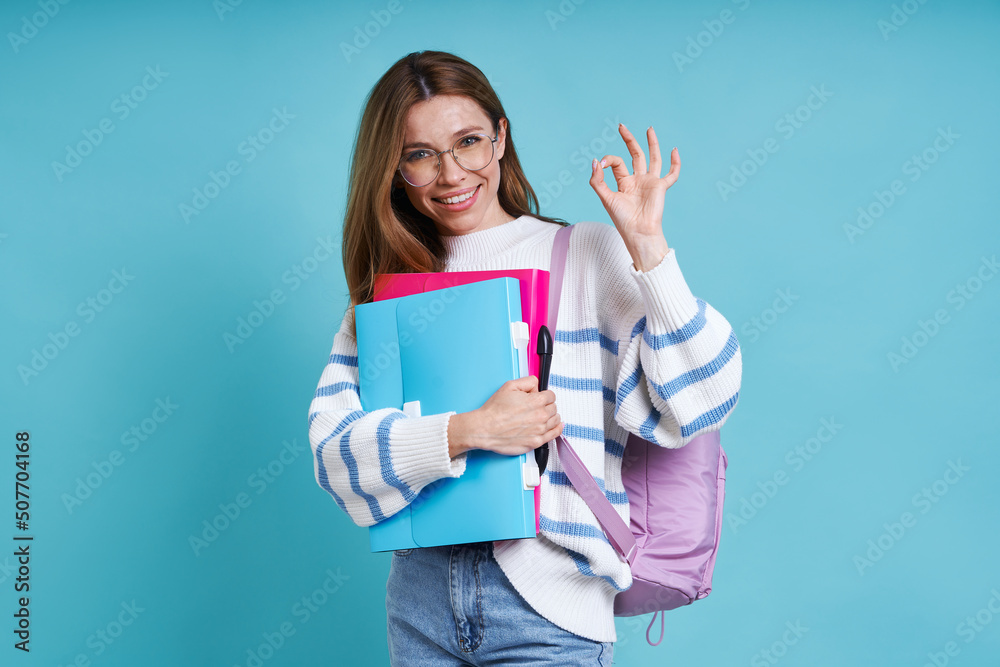 Happy female student gesturing OK sign while standing against blue background Happy female student gesturing OK sign while standing against blue background