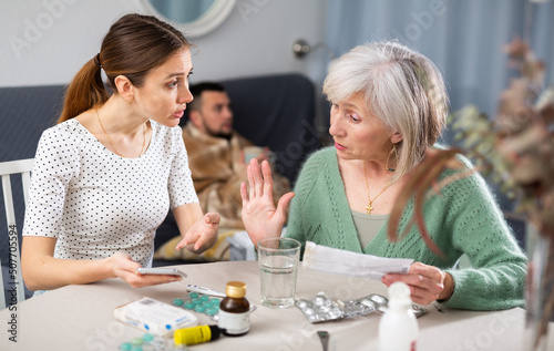Women arguing about treatment of ill man at home. Senior woman reading instruction for medicine, her daughter-in-law searching information in internet with smartphone.