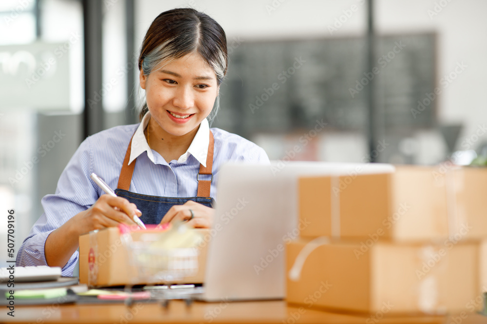 Young Asian entrepreneur SME woman checking product on stock take note with a laptop. Small business owner working at home. Check address on the parcel SME online start up freelance concept. Young Asian entrepreneur SME woman checking product on stock take note with a laptop. Small business owner working at home. Check address on the parcel SME online start up freelance concept.