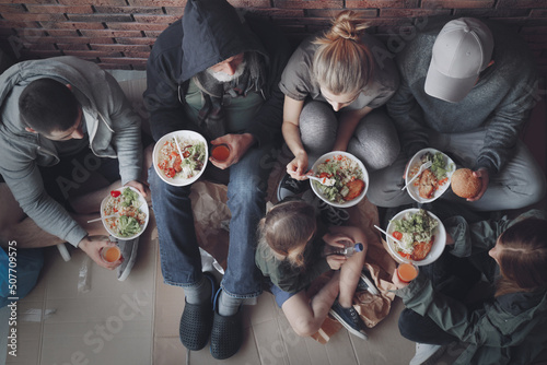 Poor people with plates of food sitting at wall indoors, view from above