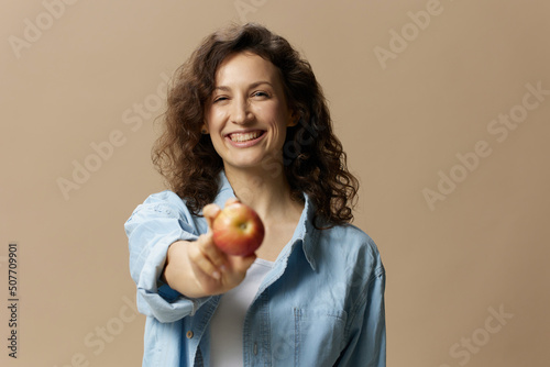 Cute cheerful happy curly beautiful female in jeans casual shirt pulls apple at camera posing isolated on over beige pastel background. Healthy food. Natural eco-friendly products concept. Copy space