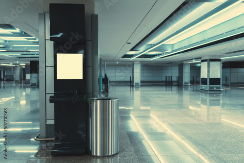 A wide-angle view of a modern opened phone booth with a payphone indoors of a contemporary airport terminal or a waiting hall of a railway station depot with a trash can next to the kiosk