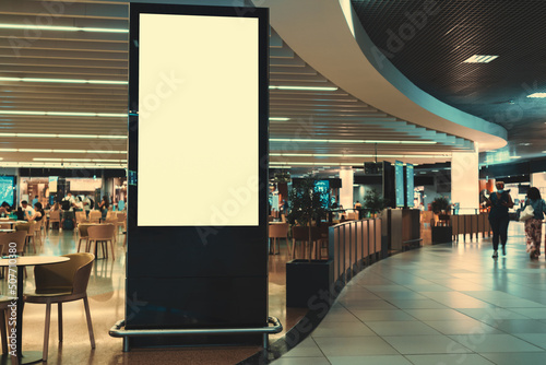 An empty vertical advertising banner mockup in front of a crowded shopping mall or an airport food court with tables and customers in the background and a copy space place on the right for an ad text