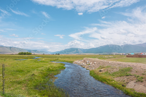 Scenic green landscape with mountain creek in sunlit grassy steppe among mountains under clouds in blue sky at changeable weather. Colorful scenery with mountain brook with clear water in bright sun.