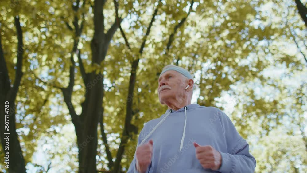 Close up of athletic fitness senior man running in sunny park. Active elderly grandfather jogging in summer forest during morning workout outdoors. Active retirement, sport healthy lifestyle concept Close up of athletic fitness senior man running in sunny park. Active elderly grandfather jogging in summer forest during morning workout outdoors. Active retirement, sport healthy lifestyle concept