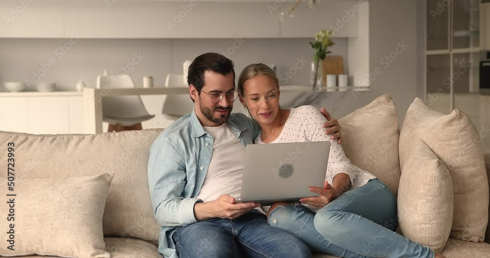 Affectionate happy family couple relaxing on cozy couch with computer on laps, enjoying watching funny photo or video content in social network, shopping or web surfing information, planning vacation. Affectionate happy family couple relaxing on cozy couch with computer on laps, enjoying watching funny photo or video content in social network, shopping or web surfing information, planning vacation.