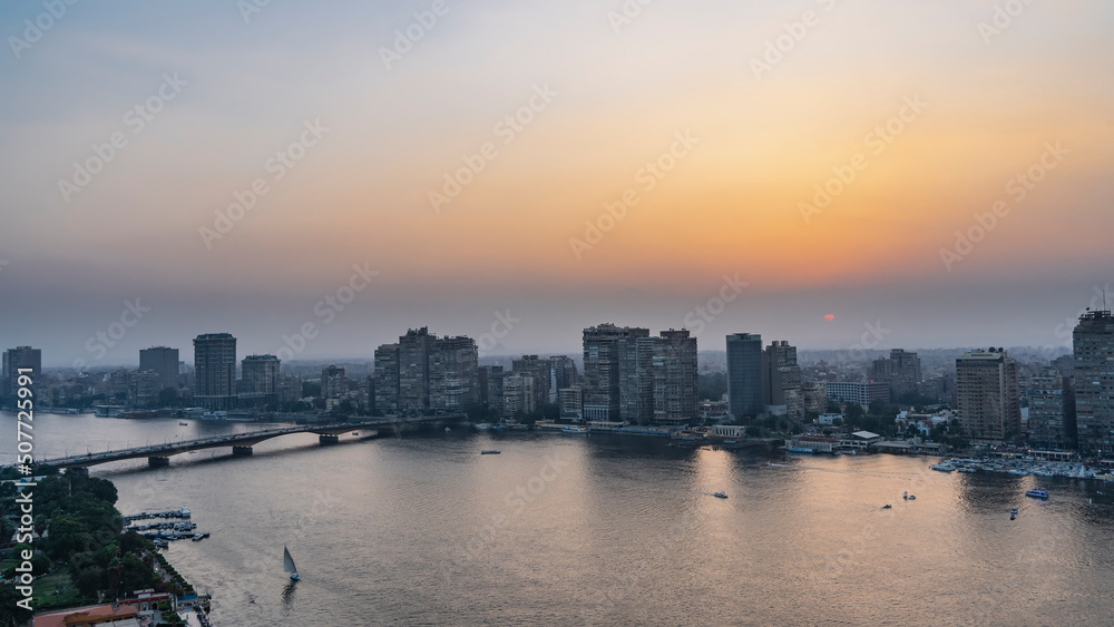 Sunset in Cairo. The sun is low in the blue-orange sky. City houses are visible on the Nile embankment. The bridge crosses the river. Reflection on the water. Aerial view. Egypt Sunset in Cairo. The sun is low in the blue-orange sky. City houses are visible on the Nile embankment. The bridge crosses the river. Reflection on the water. Aerial view. Egypt