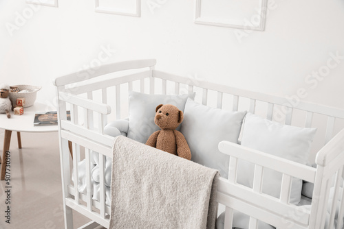 Baby crib with blanket, pillows and teddy bear near light wall photo