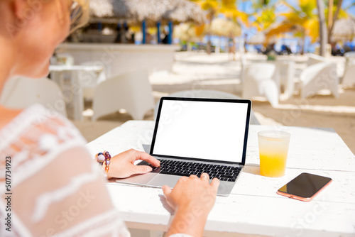 Woman working on laptop computer while sitting in tropical beach bar, empty white screen mockup