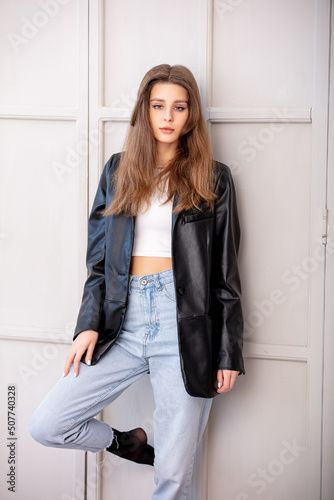 Portrait of a beautiful and young woman in jeans and a jacket standing against the wall in a bright interior.