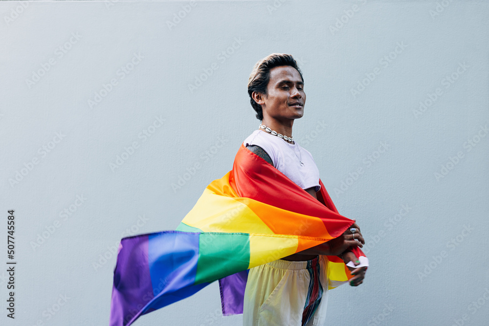 Side view of smiling male with closed eyes holding LGBT rainbow flag against grey wall