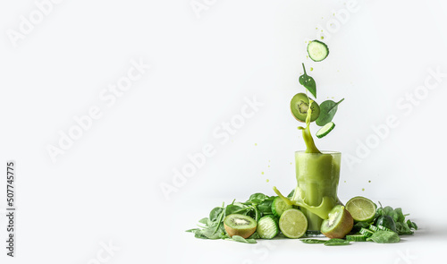 Green smoothie splashing in glass and flying ingredients: cucumber,kiwi and spinach leaves at white background with heap of green fruit and vegetable. Border. Front view.