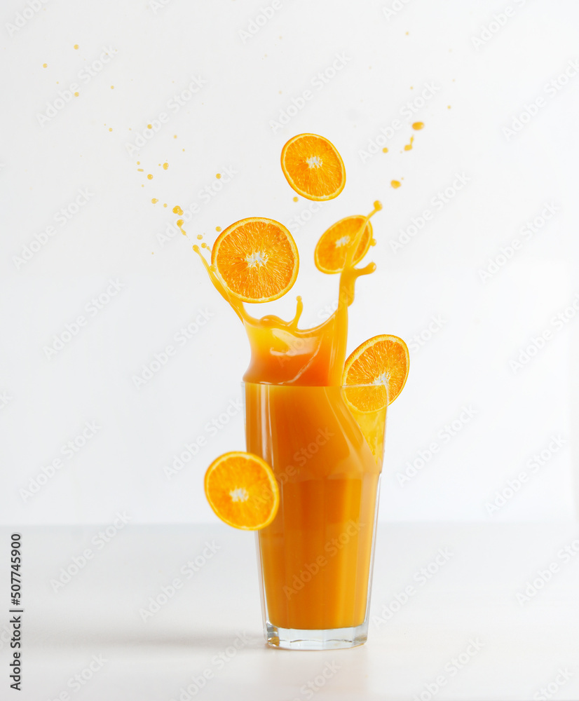Glass with splashing of orange juice and falling orange slices on table at white background. Healthy refreshing drink. Liquid motion. Front view. - obrazy, fototapety, plakaty 