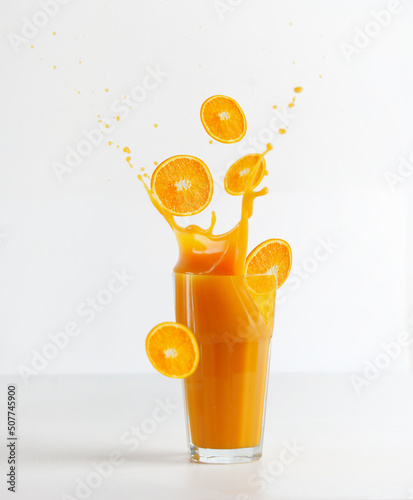 Canvas Print Glass with splashing of orange juice and falling orange slices on table at white background