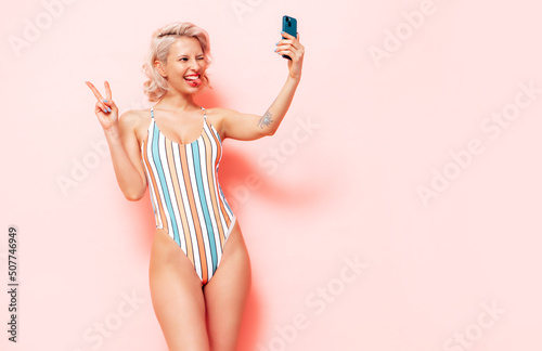 Young smiling blond model in summer swimwear colourful bathing suit. Sexy carefree woman having fun and going crazy. Female posing near pink wall in studio. Cheerful and happy. Taking selfie