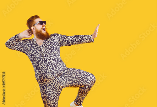 Funny eccentric overweight man dressed in leopard print pajamas dancing and fooling around on orange background. Cheerful crazy bearded fat man in sunglasses having fun at copy space. Web banner.