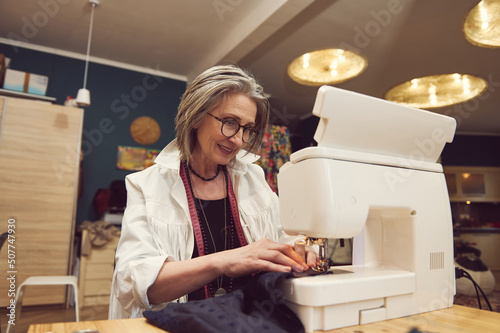 Elegant mature woman clothing designer tailor, sews clothes on sewing machine, stitches fabric, creates new clothes collection in tailoring atelier