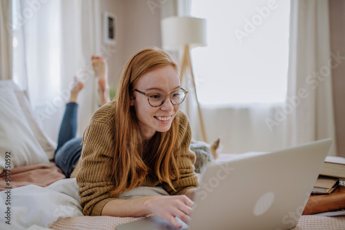 Smiling woman wearing eyeglasses using laptop lying on bed at home photo