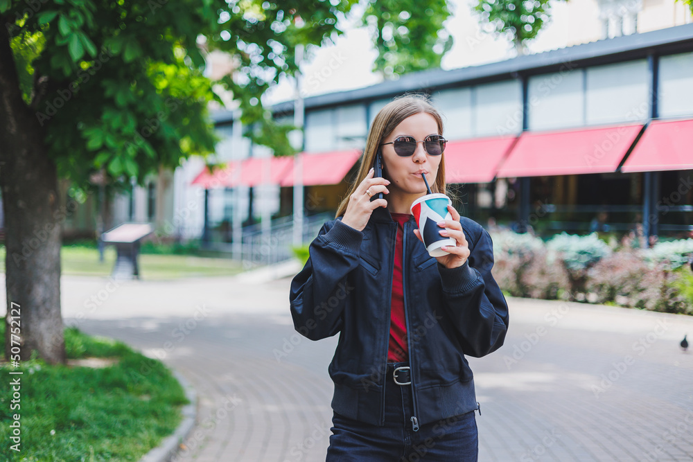 Beautiful and young girl in sunglasses writes a message on a cell phone and drinking coffee outdoors Beautiful and young girl in sunglasses writes a message on a cell phone and drinking coffee outdoors