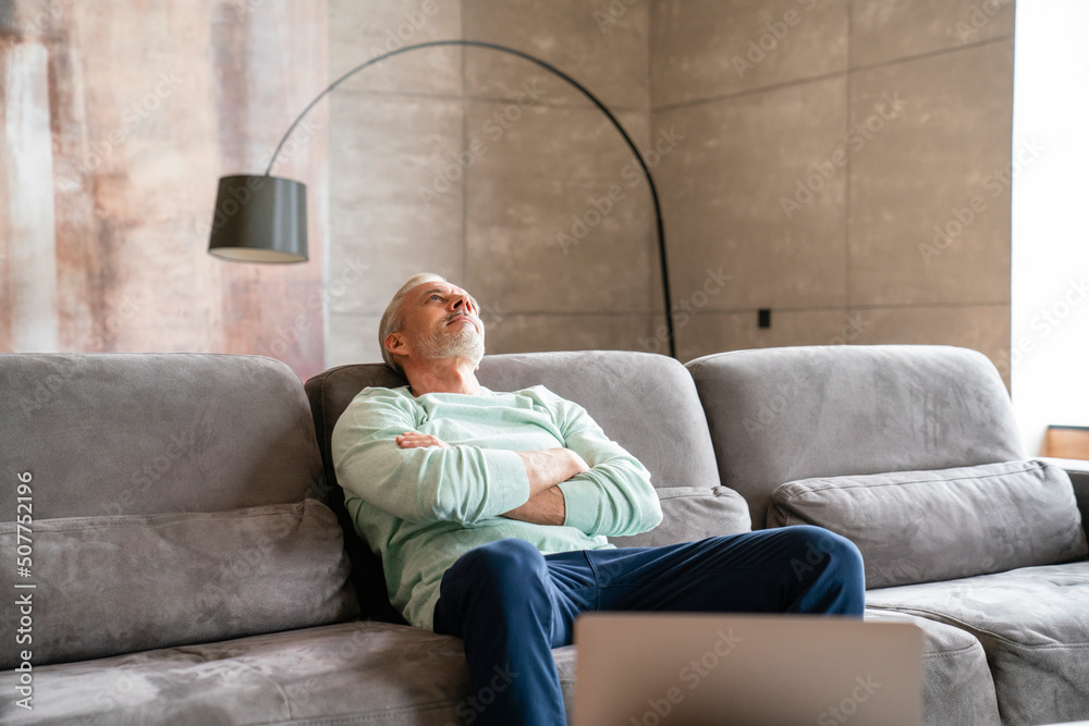 Thoughtful mature man with arms crossed sitting on sofa at home Thoughtful mature man with arms crossed sitting on sofa at home