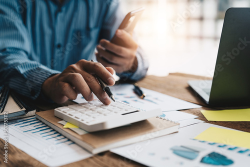 Close up of businessman or accountant hand holding mobile phone working on calculator to calculate business data, accountancy document and laptop computer at office, business concept