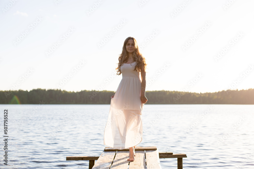 Beautiful Russian girl dressed in a white dress, walking along a wooden pier on the bank of a river or a lake