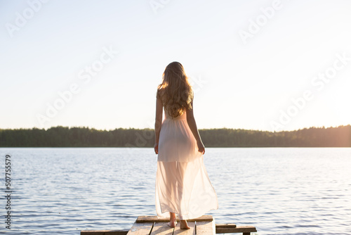 Beautiful Russian girl dressed in a white dress, walking along a wooden pier on the bank of a river or a lake