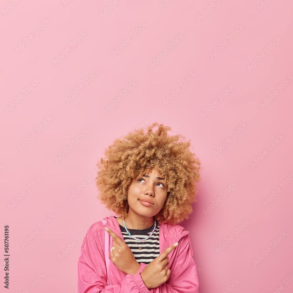 Hesitant curly haired woman points index fingers sideways dressed in jacket chooses between two options focused above thoughtfully isolated over pink background blank space for advertisement