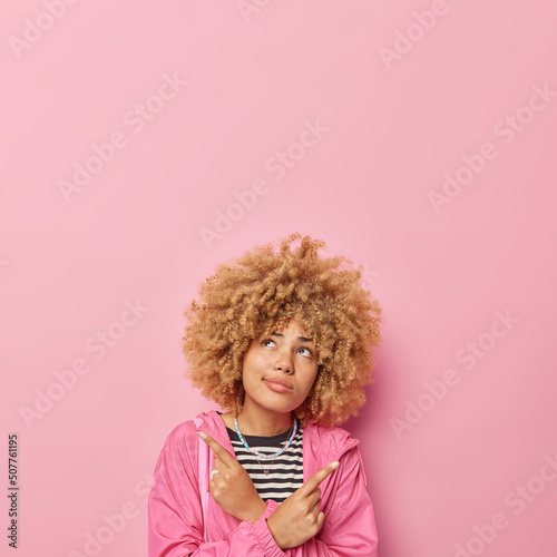 Hesitant curly haired woman points index fingers sideways dressed in jacket chooses between two options focused above thoughtfully isolated over pink background blank space for advertisement