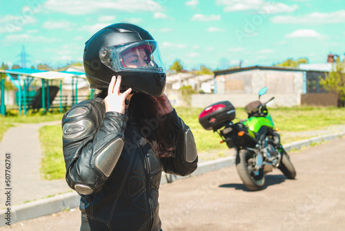 A girl in a protective motorcycle leather suit puts on a helmet against the backdrop of a motorcycle.