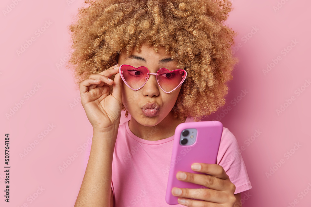 Pretty curly haired young woman keeps lips folded looks at smartphone checks message from boyfriend wears trendy sunglasses casual t shirt isolated over pink background uses mobile phone app