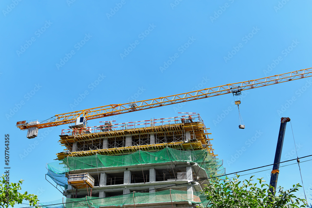 Naklejka premium A yellow tower crane lifts a cargo on a building construction site with a green mesh against a clear blue sky